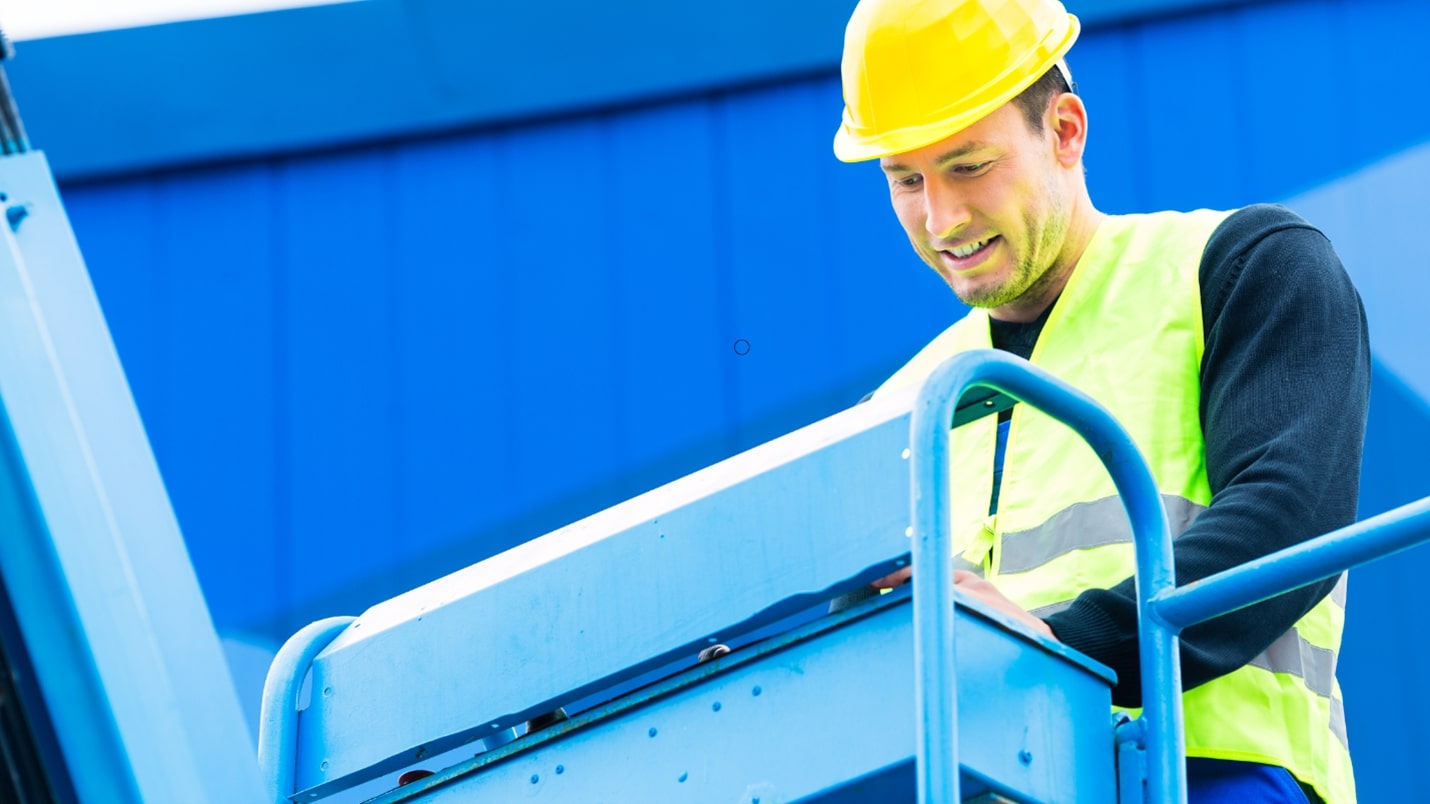 A man in a yellow hard hat and safety vest is doing maintenance of a Morn lift at a construction site.