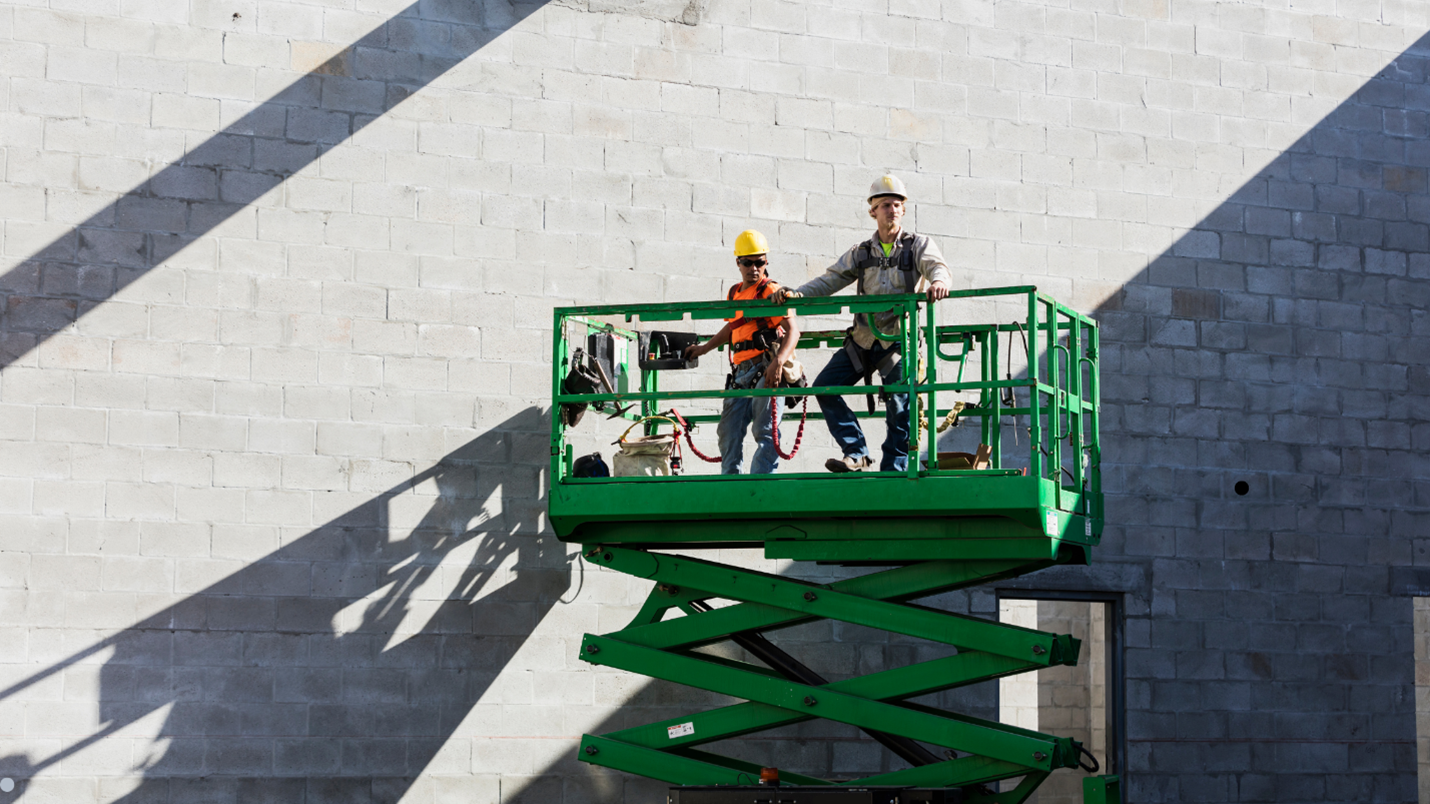 Two men on a morn green scissor lift in front of a building, working on maintenance tasks.